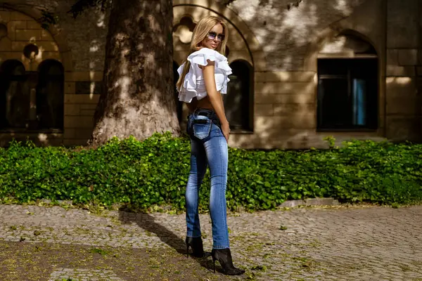 Çok ince vücutlu seksi sarışın manken, güneşli bir yaz gününde şehirde dar kotlar ve dalgalı beyaz bir bluzla şehvetle poz veriyor. Kot pantolon moda..