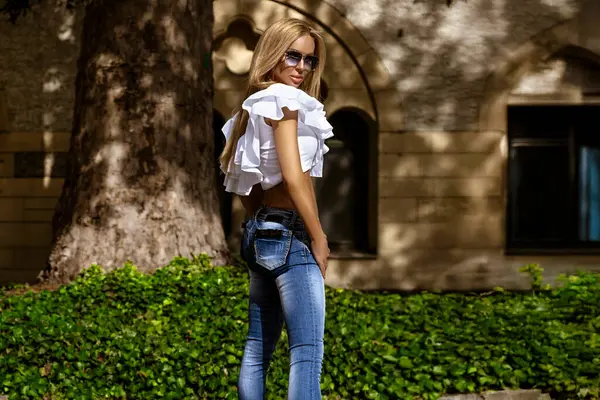Çok ince vücutlu seksi sarışın manken, güneşli bir yaz gününde şehirde dar kotlar ve dalgalı beyaz bir bluzla şehvetle poz veriyor. Kot pantolon moda..