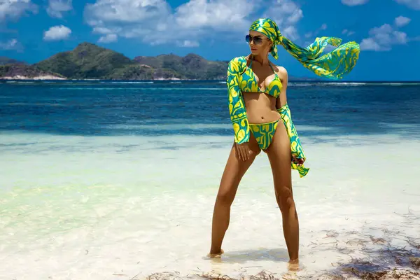 Tropikal yeşil bikinili şık bir kadın ve arka planda turkuaz su ve tekneyle Seyşeller 'deki güneşli egzotik plajlarda poz veren eşarp.
