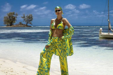 Seksi bikinili, eşarp ve plaj pantolonu giyen şık bir kadın Seyşeller 'deki kumlu sahilde poz veriyor. Plaj kıyafeti modası.