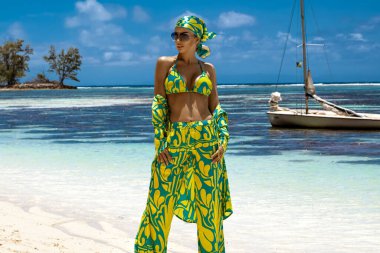 Seksi bikinili, eşarp ve plaj pantolonu giyen şık bir kadın Seyşeller 'deki kumlu sahilde poz veriyor. Plaj kıyafeti modası.