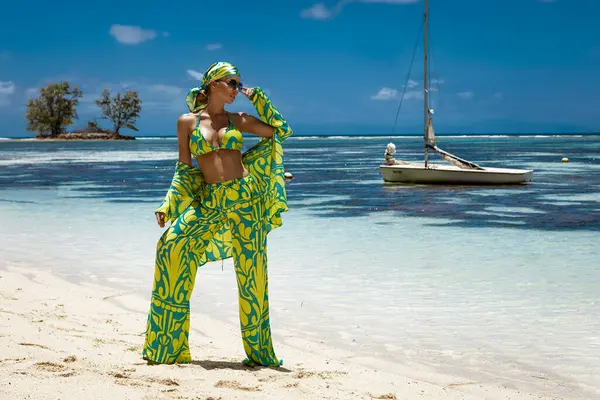 Seksi bikinili, eşarp ve plaj pantolonu giyen şık bir kadın Seyşeller 'deki kumlu sahilde poz veriyor. Plaj kıyafeti modası.