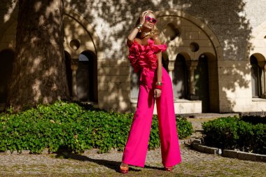 Kalın fuşya tüylü ceketi ve fırfırlı üst ve topuklu ayakkabılarıyla yaz güneşinde açık havada poz veren şık bir manken, abartılı moda ve lüks sokak tarzını ifade ediyor..