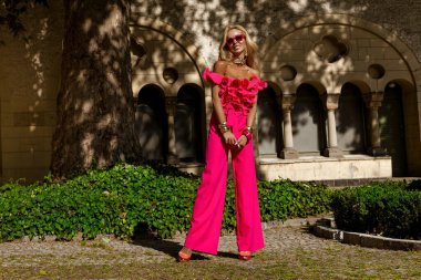 Kalın fuşya tüylü ceketi ve fırfırlı üst ve topuklu ayakkabılarıyla yaz güneşinde açık havada poz veren şık bir manken, abartılı moda ve lüks sokak tarzını ifade ediyor..