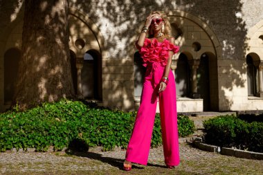 Kalın fuşya tüylü ceketi ve fırfırlı üst ve topuklu ayakkabılarıyla yaz güneşinde açık havada poz veren şık bir manken, abartılı moda ve lüks sokak tarzını ifade ediyor..