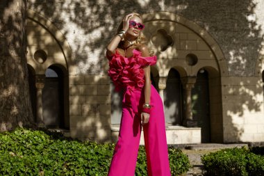Kalın fuşya tüylü ceketi ve fırfırlı üst ve topuklu ayakkabılarıyla yaz güneşinde açık havada poz veren şık bir manken, abartılı moda ve lüks sokak tarzını ifade ediyor..