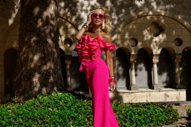 Kalın fuşya tüylü ceketi ve fırfırlı üst ve topuklu ayakkabılarıyla yaz güneşinde açık havada poz veren şık bir manken, abartılı moda ve lüks sokak tarzını ifade ediyor..