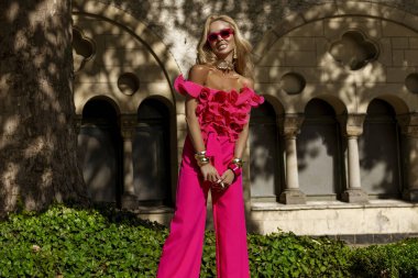 Kalın fuşya tüylü ceketi ve fırfırlı üst ve topuklu ayakkabılarıyla yaz güneşinde açık havada poz veren şık bir manken, abartılı moda ve lüks sokak tarzını ifade ediyor..