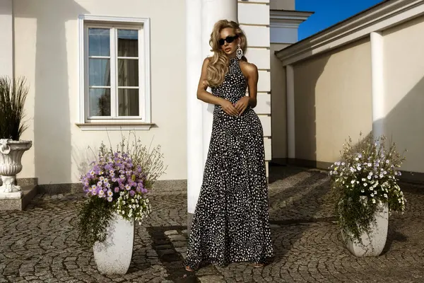 Siyah benekli Maxi elbiseli zarif bir kadın lüks villanın önünde poz veriyor, yaz mankeni güneş gözlüğü takıyor ve küpe takıyor.