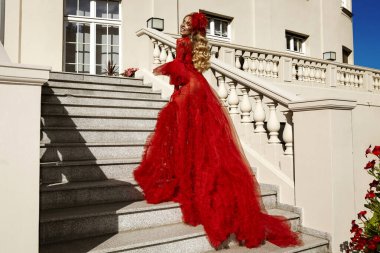 Lüks kırmızı elbisesi içinde zarif bir kadın. Merdivenlerde tüy detaylarıyla poz veriyor. Yaz günü gece elbisesiyle fotoşoplar çekiyor.