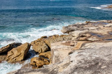 Okyanus dalgaları güneşli bir günde kıyı boyunca dokulu kum taşı oluşumlarına çarpar. Kraliyet Ulusal Parkı, NSW, Avustralya