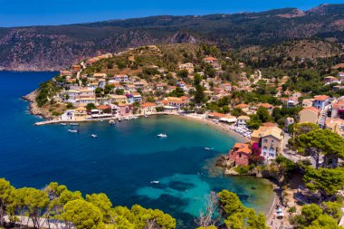 Assos 'un havadan görünüşü, Kefalonia. Turkuaz sularla çevrili bir yarımadada yuva yapmış güzel bir köy. Renkli evler, küçük plajlar ve körfezdeki tekneler. Arkaplanda yemyeşil tepeler ve dağlar var..