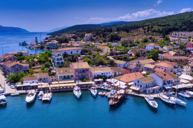 Aerial view of Fiskardo, Kefalonia. Picturesque village nestled on a peninsula, surrounded by turquoise waters. Colorful houses, small beach, and boats in the bay. Lush green hills and mountains in the background clipart