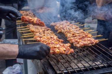 Açık ateşte pişen et şişleriyle dolu bir ızgara. Birisi eldiven giyerek ızgarayla ilgilenirken et kahverengiler gibi duman yükselir. Bu sahnede geleneksel bir barbekü yapılıyor..