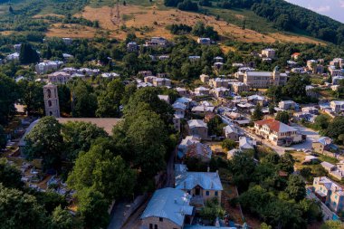 An aerial view of Nymfaio, a picturesque mountain village in Greece, surrounded by lush green forests and hills. The village's traditional stone houses with metal roofs are nestled into the scenic landscape. clipart