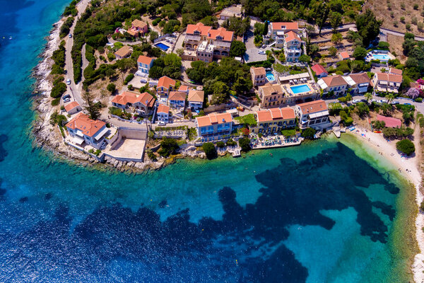 Вид с воздуха на Фискардо, Кефалония. Живописная деревня, расположенная на полуострове, окруженная бирюзовыми водами. Красочные дома, небольшой пляж и лодки в заливе. Пышные зеленые холмы и горы на заднем плане