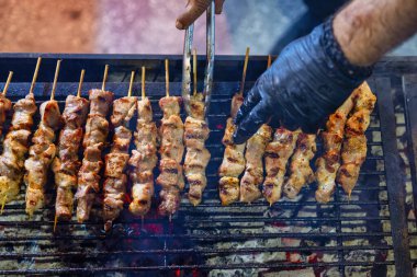 Açık ateşte pişen et şişleriyle dolu bir ızgara. Birisi eldiven giyerek ızgarayla ilgilenirken et kahverengiler gibi duman yükselir. Bu sahnede geleneksel bir barbekü var. Greek Souvlaki