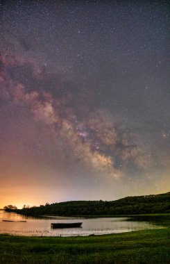Samanyolu Kerkini Gölü 'nün durgun suları üzerinde parlak bir şekilde parıldıyor. Yalnız bir tekne, doğanın barışçıl kucaklamalarıyla nazikçe sürüklenir. Yıldızların yansıması rüya gibi atmosferi güçlendirir.