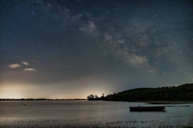 Samanyolu Kerkini Gölü 'nün durgun suları üzerinde parlak bir şekilde parıldıyor. Yalnız bir tekne, doğanın barışçıl kucaklamalarıyla nazikçe sürüklenir. Yıldızların yansıması rüya gibi atmosferi güçlendirir.