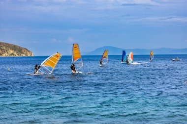 Limni, Evia, Yunanistan 21 Haziran 2025: Rüzgâr sörfçüleri renkli yelkenlerle denizde süzülerek yaz sporlarının enerji ve güzelliklerini manzaralı bir kıyı manzarasında yakalıyorlar.