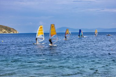 Limni, Evia, Yunanistan 21 Haziran 2025: Rüzgâr sörfçüleri renkli yelkenlerle denizde süzülerek yaz sporlarının enerji ve güzelliklerini manzaralı bir kıyı manzarasında yakalıyorlar.