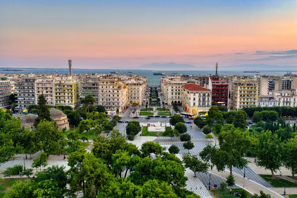 Selanik, Yunanistan 3 Ağustos 2025. Pembe gökyüzü, şehir mimarisi, termaik körfezdeki gemiler ve yeşil park alanlarıyla şehir merkezinin hava aracı görüntüsü.