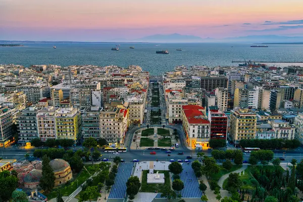 Selanik, Yunanistan 3 Ağustos 2025. Pembe gökyüzü, şehir mimarisi, termaik körfezdeki gemiler ve yeşil park alanlarıyla şehir merkezinin hava aracı görüntüsü.