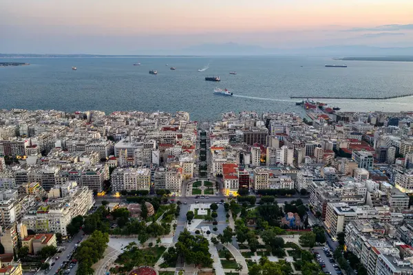 Selanik, Yunanistan 3 Ağustos 2025. Pembe gökyüzü, şehir mimarisi, termaik körfezdeki gemiler ve yeşil park alanlarıyla şehir merkezinin hava aracı görüntüsü.