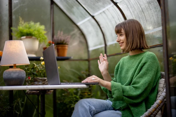 年轻女人在笔记本电脑上工作 一边坐在圆桌边聊天 一边坐在温室里 后院种着植物和鲜花 在舒适的环境中工作 — 图库照片
