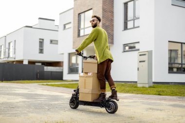 Adam elektrikli scooter kullanıyor, yerleşim bölgesinde caddede karton kutular dağıtıyor. Sürdürülebilirlik kavramı, dağıtım ve çevre dostu modern yaşam tarzı