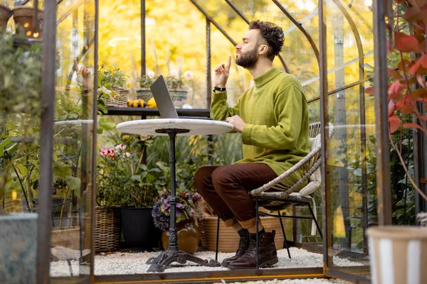 裏庭の植物や花とガラスの家の丸いテーブルに座っている間 男はノートパソコンで動作します 自然の中で居心地の良い雰囲気で自宅から仕事 — ストック写真