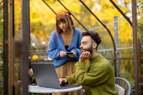 ガラスの家の丸いテーブルに植物や花がある裏庭でノートパソコンで仕事をしながら 男と女は会話をしています 自然の中で居心地の良い雰囲気で自宅から仕事 — ストック写真