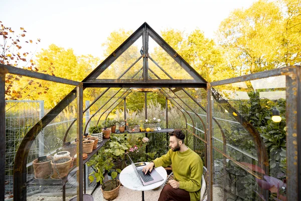 男人在笔记本电脑上工作 而坐在圆桌旁边的温室里 后院种着植物和鲜花 在舒适的环境中工作 — 图库照片
