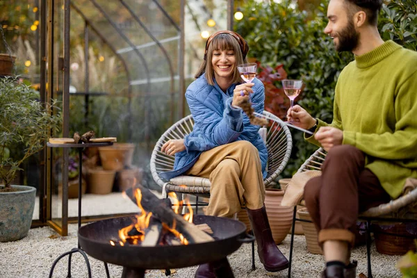 若いスタイリッシュなカップルは食べ物を焼くと一緒に火のそばに座っている間に暖まる 庭の居心地の良い雰囲気で秋の夜の時間を過ごす — ストック写真