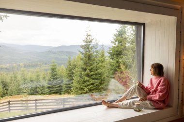Kadın pencere pervazında telefonla oturur ve doğadaki ahşap evde dinlenirken dağların manzarasından zevk alır. Eğlence ve doğa kavramına kaçmak