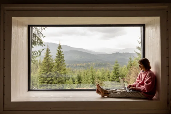 Kadın Dağların Manzarası Harika Olan Bir Pencerede Otururken Dizüstü Bilgisayarla — Stok fotoğraf