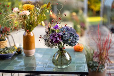 Arka bahçedeki masanın üstünde vazoda duran bir buket güzel çiçek ve arka planda sera.