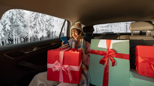身着节日服装的年轻女子坐在后座上开车 带着包装精美的圣诞礼物和电话 背景上的雪景 广角镜 — 图库照片