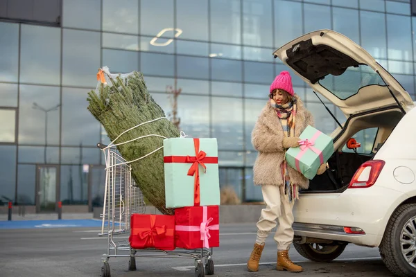 Young woman packing christmas presents and tree into car, standing with trolley at parking lot of a mall. Cheerful girl having festive shopping in anticipation of the winter holidays