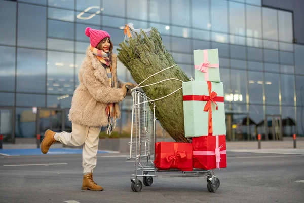 一个女人把满是礼物和圣诞树的购物车推到外面的购物中心前 寒假购物概念 — 图库照片
