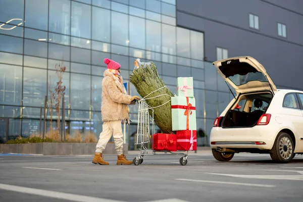 一个女人把装满礼物和圣诞树的购物车推到商场停车场 寒假购物概念 — 图库照片