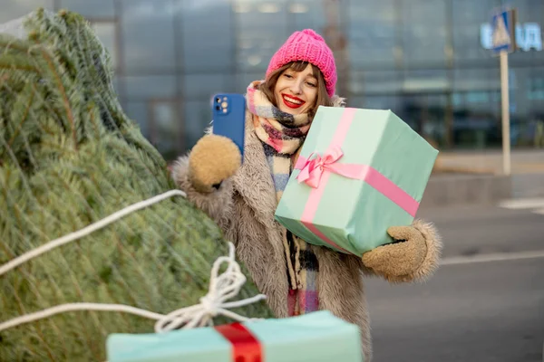 年轻快乐的女人站在商场前的购物车旁 满载着礼物和圣诞树 一边打电话一边聊天 — 图库照片