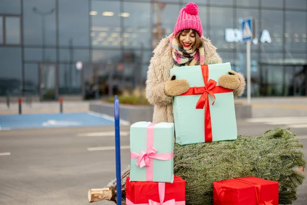 屋外のモールの近くにギフトやクリスマスツリーでいっぱいのショッピングトロリーで立って冬服の若い女性の肖像画 冬休みショッピングのコンセプト — ストック写真