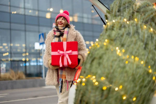 冬服の若い陽気な女性が前にショッピングモールやクリスマスツリーの近くのギフトボックスで立っています 冬休みの買い物のコンセプト — ストック写真
