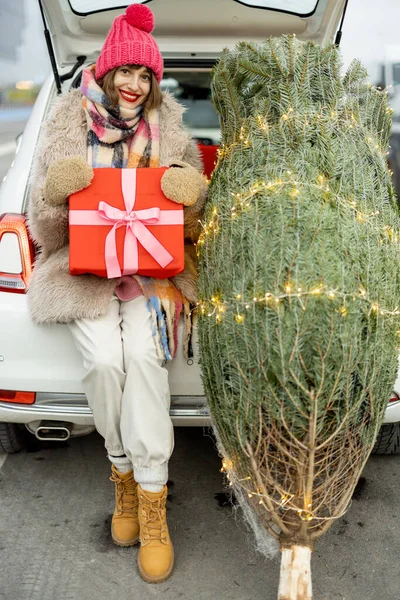 一个带着礼品盒的年轻而快乐的女人的画像 她刚买了一棵圣诞树 坐在商场停车场的汽车行李箱旁 寒假购物的概念 — 图库照片