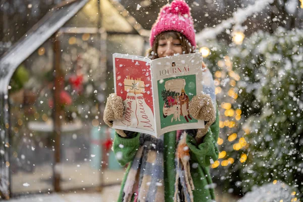 年轻女人在为寒假装饰的雪地后院里读一些关于新年主题的杂志 出版新年假期的概念 — 图库照片
