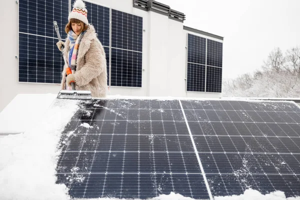 Woman cleans solar panels from snow to produce power in winter on the roof of her house. Energy independence and sustainability concept