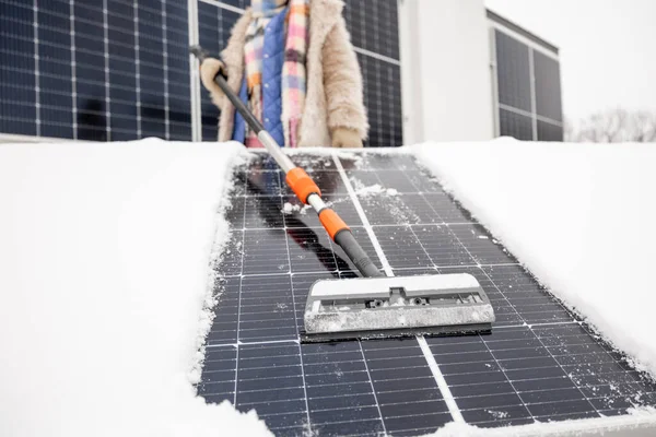 女性は家の屋根の上で冬に発電するために雪から太陽電池パネルをきれいにします エネルギー自立と持続可能性の概念 綿棒に焦点を当てた底からの眺め — ストック写真