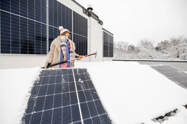 Woman cleans solar panels from snow to produce power in winter on the roof of her house on nature. Energy independence and sustainability concept. Wide angle view