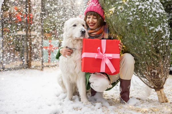 雪の上の美しい装飾された庭で彼女の愛らしい犬とクリスマスツリーとギフトボックスの抱擁を持つ女性の肖像画 幸せな冬の休日と魔法の概念 — ストック写真